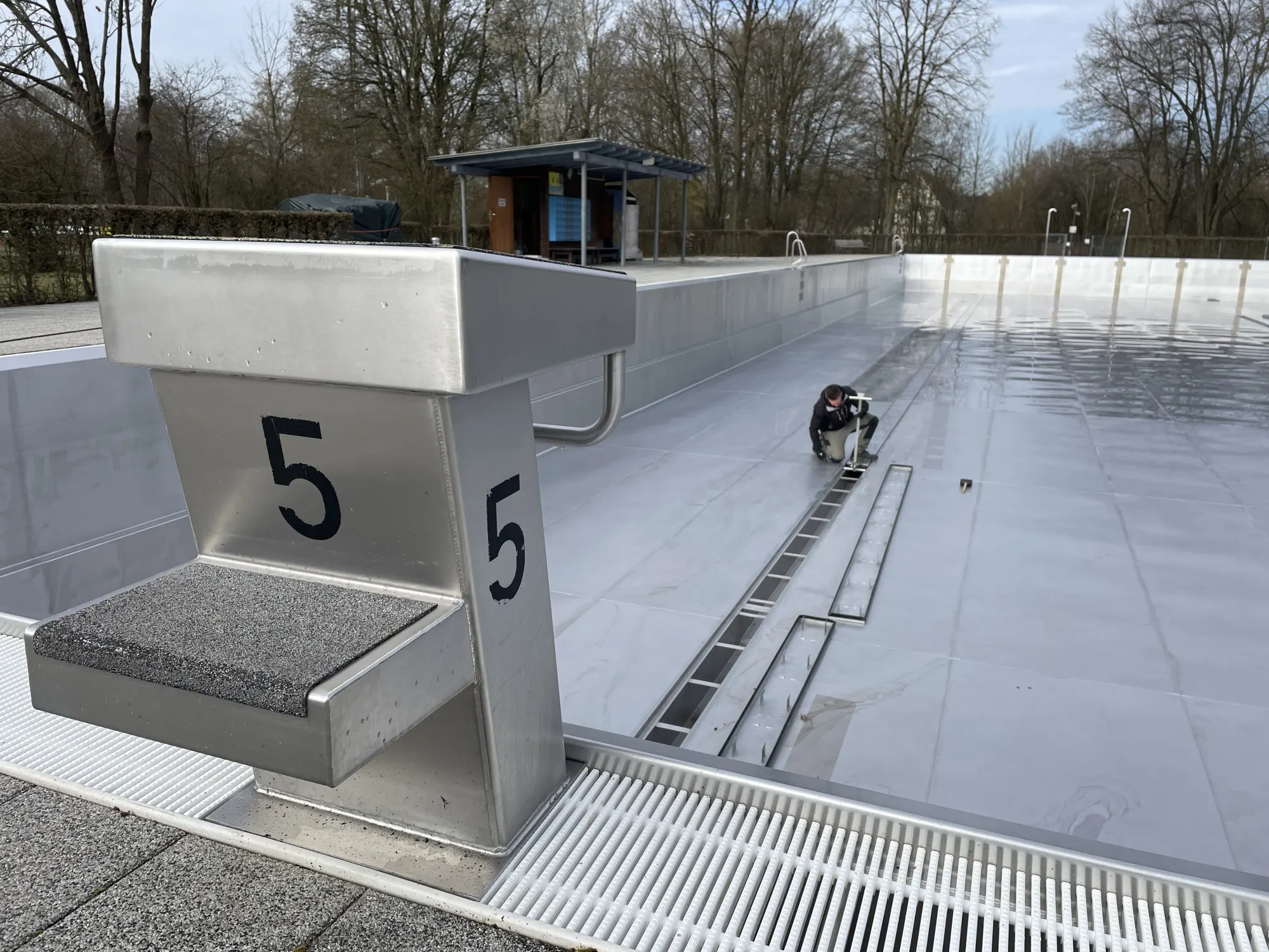 Leeres Freibad mit nummerierten Startblöcken, Person arbeitet am Beckenrand, Bäume im Hintergrund