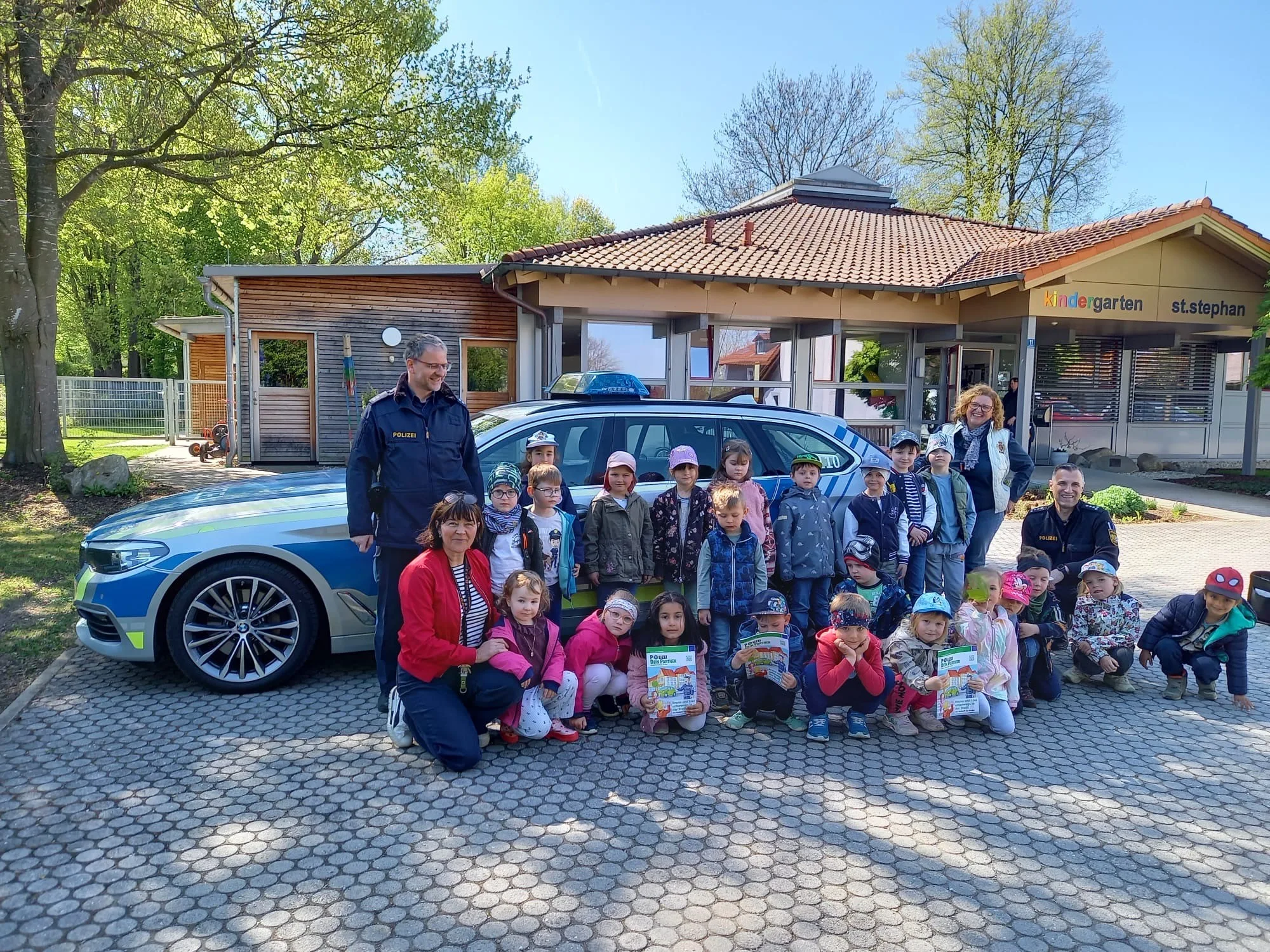 Polizeibeamte posieren mit Kindergartenkindern vor Polizeifahrzeug auf gepflastertem Hof vor Kindergartengebäude