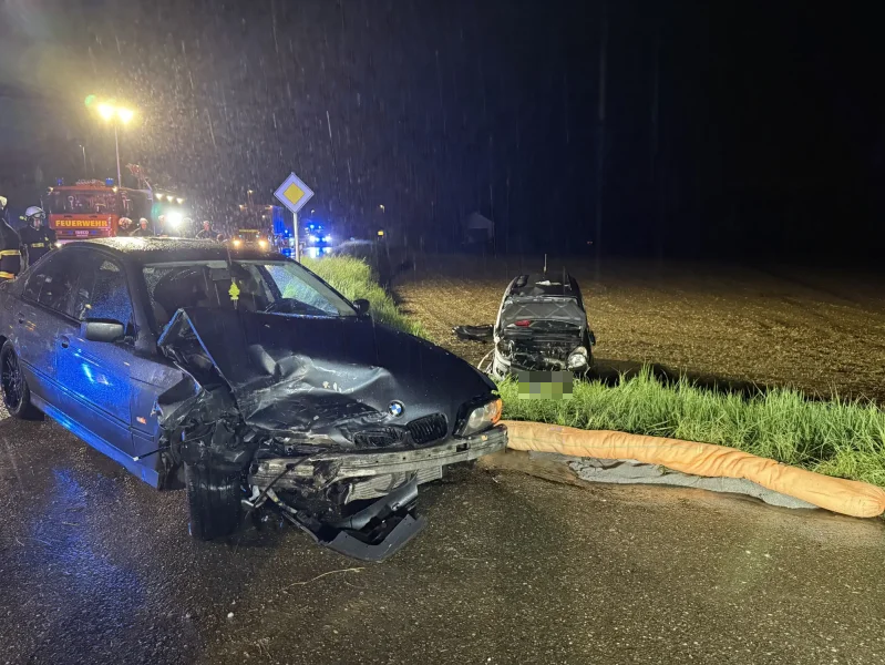 Schwer beschädigter BMW nach Verkehrsunfall bei Regen, Feuerwehrfahrzeuge im Hintergrund