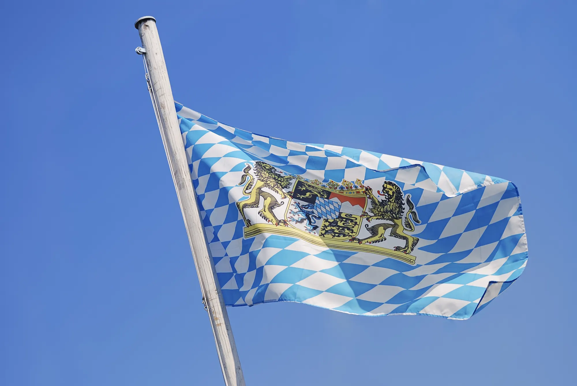 Bayerische Flagge mit blau-weißen Rauten und Wappen weht am Mast vor blauem Himmel
