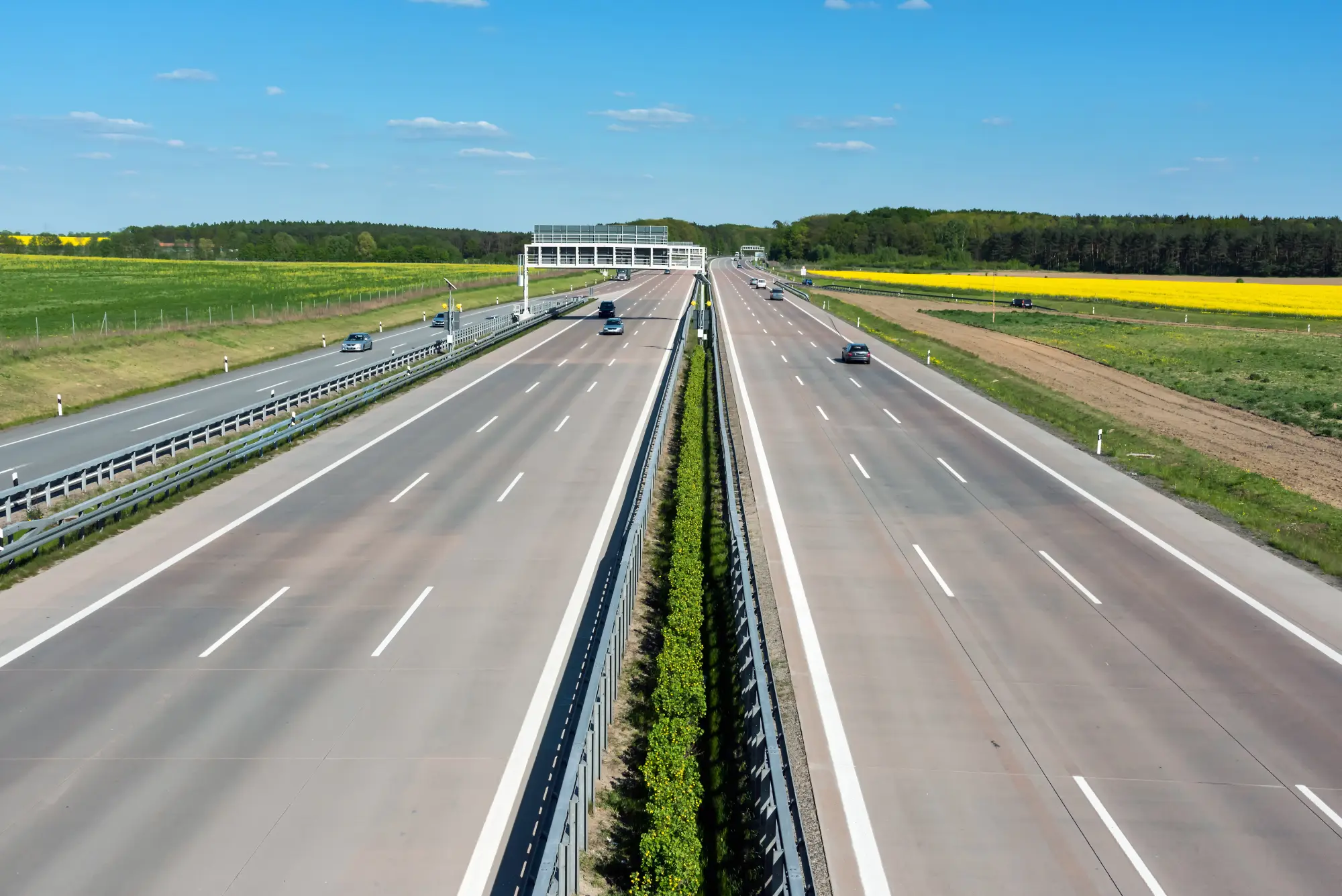 Mehrspurige Autobahn mit grünem Mittelstreifen führt durch ländliche Landschaft mit Feldern und Wald