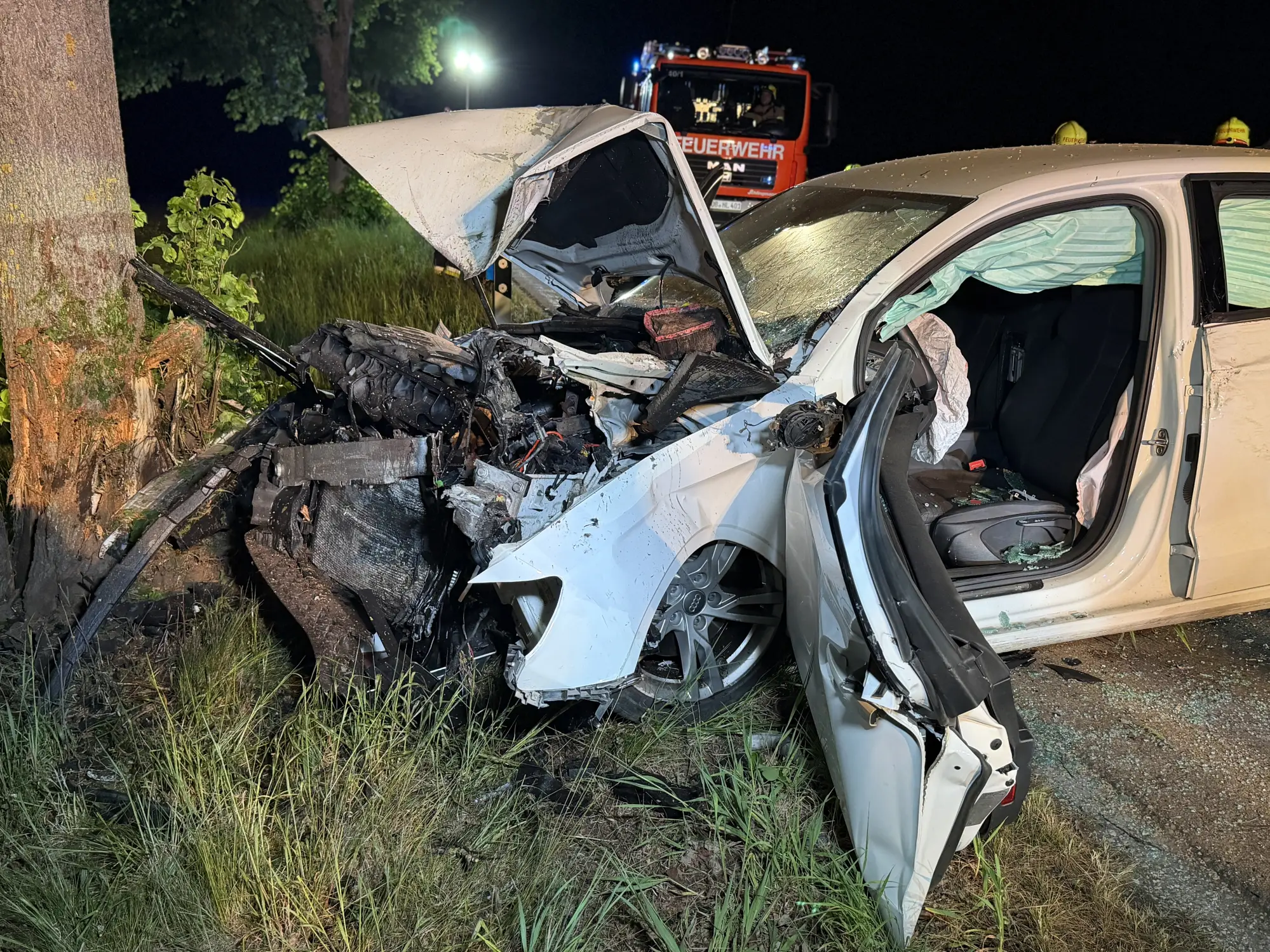 Schwer beschädigtes Auto nach Verkehrsunfall, Feuerwehrfahrzeug im Hintergrund, Fahrzeug gegen Baum geprallt