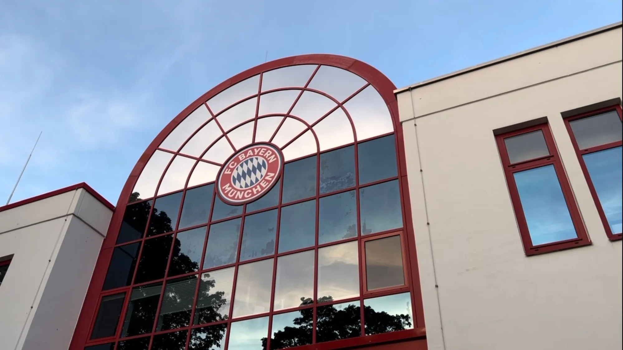 Gebäude mit großer Glasfront und FC Bayern München Logo, rote Fensterrahmen, blauer Himmel im Hintergrund