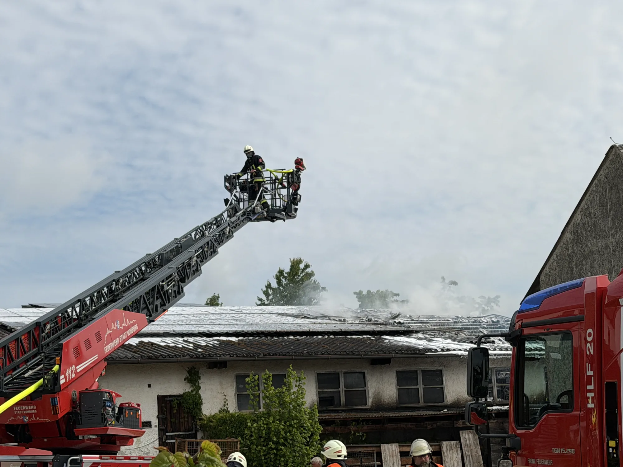 Feuerwehr-Drehleiter im Einsatz bei Lagerhallenbrand, Rauch steigt vom Dach auf, mehrere Einsatzfahrzeuge vor Ort