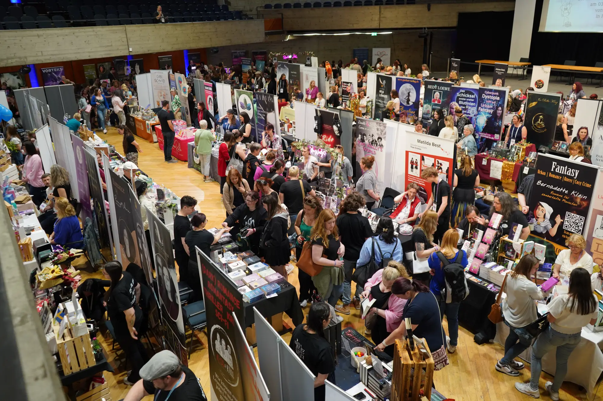 Übersicht einer gut besuchten Buchmesse mit vielen Ständen und Besuchern in einer großen Halle